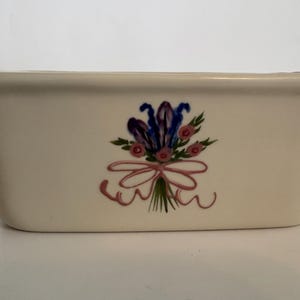 May include: Cream-colored rectangular ceramic container with a floral bouquet design. The bouquet features purple, blue, and pink flowers tied with a pink ribbon. The container has a slightly curved shape and is likely for decorative or functional use.