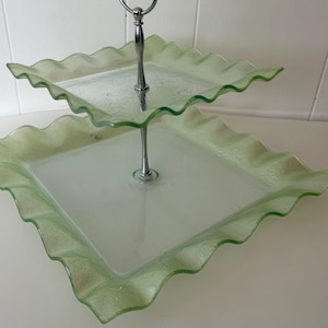 Peut inclure: Un présentoir à deux niveaux composé de deux plateaux carrés en verre vert clair aux bords ondulés et texturés. Les niveaux sont reliés par une tige centrale en métal argenté poli, surmontée d'une poignée circulaire.