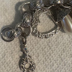 May include: A silver charm bracelet with various charms, including a heart-shaped charm, a faux pearl, and a decorative pendant. The bracelet has a mix of textures and designs, with intricate detailing on the charms.