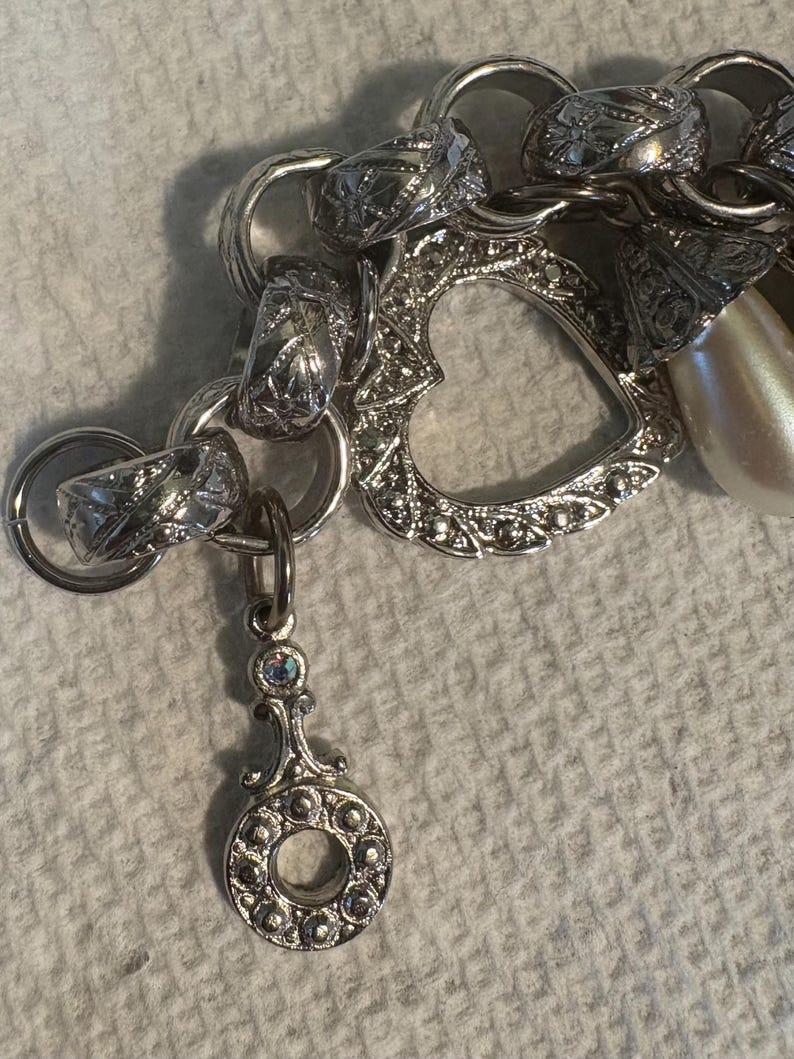 May include: A silver charm bracelet with various charms, including a heart-shaped charm, a faux pearl, and a decorative pendant. The bracelet has a mix of textures and designs, with intricate detailing on the charms.