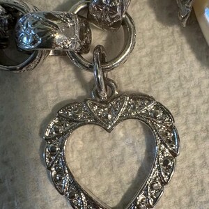 May include: A silver-toned heart-shaped pendant with an open center, attached to a chain. The pendant has a detailed, ornate border. The chain links are also intricately designed, showcasing a vintage aesthetic.