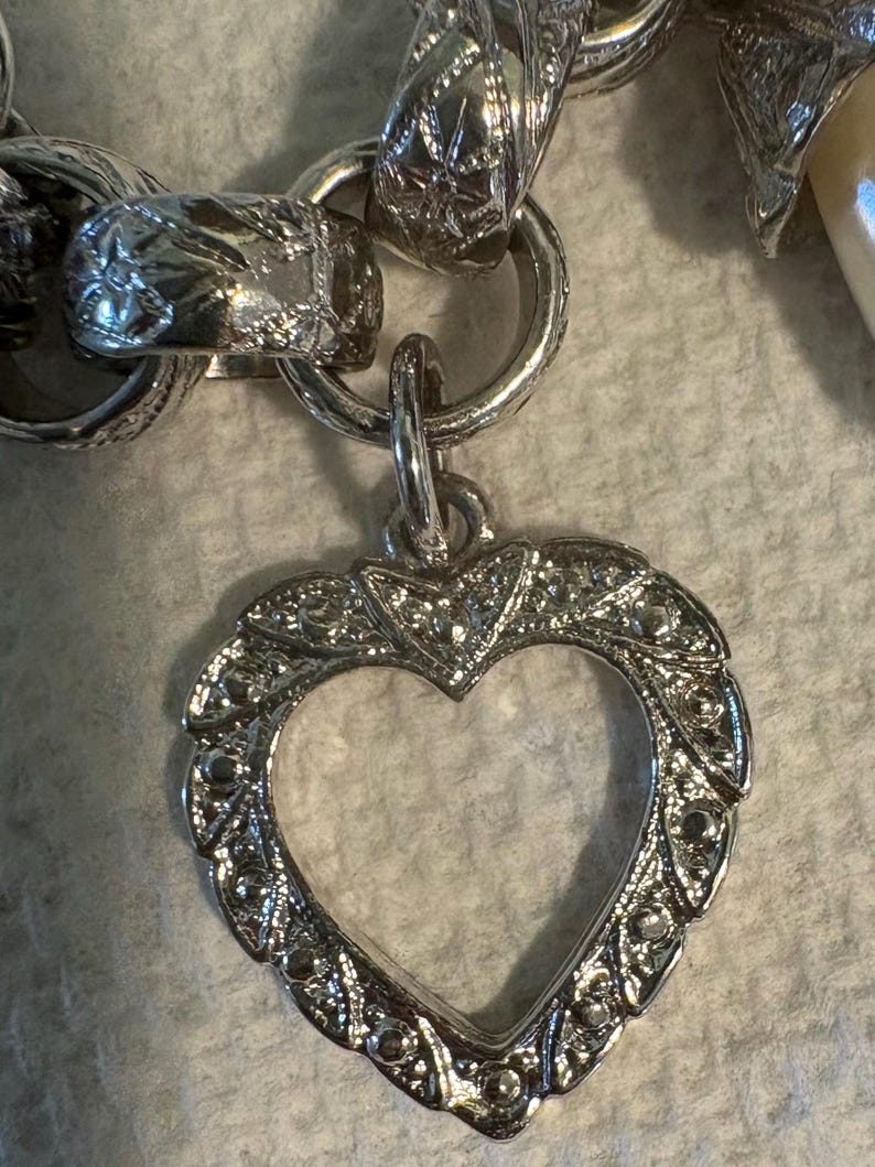 May include: A silver-toned heart-shaped pendant with an open center, attached to a chain. The pendant has a detailed, ornate border. The chain links are also intricately designed, showcasing a vintage aesthetic.