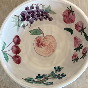 May include: White ceramic bowl with hand-painted fruit illustrations. The bowl features images of grapes, strawberries, cherries, a peach, and blueberries. The fruit is painted in shades of red, purple, and green, creating a colorful and decorative design.