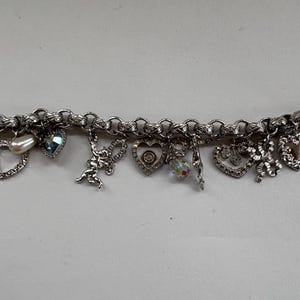 May include: Silver charm bracelet featuring various charms, including hearts, a pearl, a blue gemstone, a cupid, and a crystal. The bracelet has a lobster clasp closure and is designed with a chain link style.