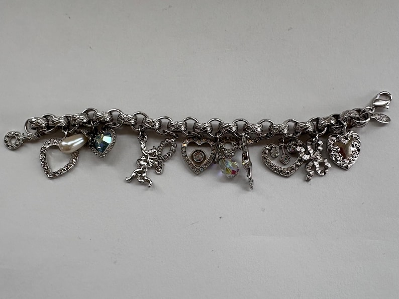 May include: Silver charm bracelet featuring various charms, including hearts, a pearl, a blue gemstone, a cupid, and a crystal. The bracelet has a lobster clasp closure and is designed with a chain link style.