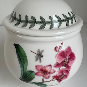May include: A white ceramic lidded jar featuring a botanical illustration of pink orchid flowers and a dragonfly on the body. The lid is decorated with a green leaf vine pattern around the edge. The jar has a rounded shape and a smooth finish.