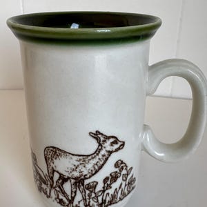 May include: A ceramic mug with a cream-colored body and a dark green rim. The exterior features a brown line-art illustration of a deer standing among plants and flowers. The mug has a curved handle on the right side and a smooth, cylindrical shape.