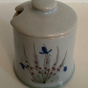 Może przedstawiać: Jasnoniebieski ceramiczny słoik z pokrywką i małym uchwytem. Słoik jest ozdobiony kwiatowym wzorem w kolorze niebieskim, czerwonym i zielonym. Słoik jest cylindryczny i ma mały guzik na górze pokrywki.