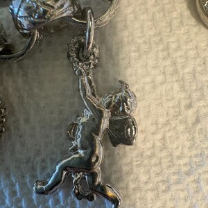 May include: Silver charm bracelet with a detailed cherub pendant. The cherub is holding a wreath above its head. The bracelet links are also silver and have ornate designs. The background is a light, textured surface.