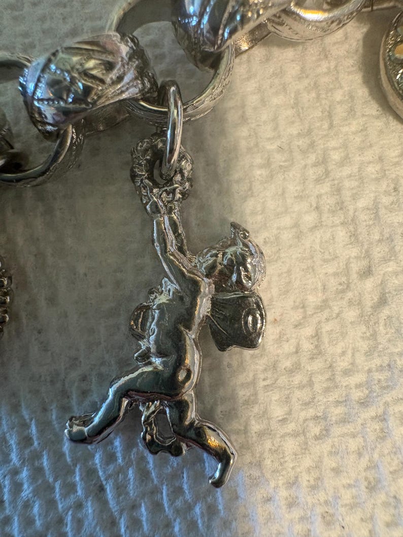 May include: Silver charm bracelet with a detailed cherub pendant. The cherub is holding a wreath above its head. The bracelet links are also silver and have ornate designs. The background is a light, textured surface.