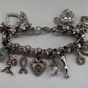 May include: A silver charm bracelet with various charms, including a pearl, heart, bow, and teardrop-shaped charms. Some charms have clear and blue crystals. The bracelet is made of a thick chain and has a clasp closure.