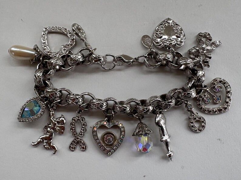 May include: A silver charm bracelet with various charms, including a pearl, heart, bow, and teardrop-shaped charms. Some charms have clear and blue crystals. The bracelet is made of a thick chain and has a clasp closure.