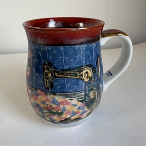 May include: A ceramic mug featuring a dark red rim and a blue body decorated with a vintage sewing machine illustration over a colorful patchwork quilt pattern. The handle is white, and the text "ORIGIN JAPAN" is printed on the inner handle base.