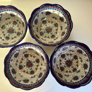 Può includere: Quattro ciotole in ceramica con bordi smerlati, caratterizzate da un interno color crema decorato con un motivo ripetitivo di fiori blu e rossi, rifinite con un bordo blu scuro solido e una sottile linea rossa tratteggiata.