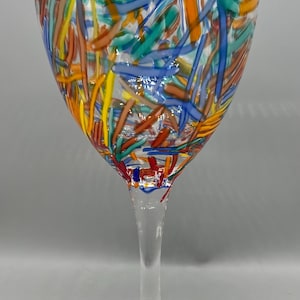 May include: A clear wine glass with a colorful abstract design. The glass features a long stem and a wide bowl, decorated with streaks of blue, yellow, orange, and brown. The rim has a silver trim.