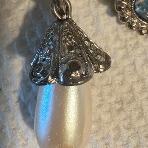 May include: Close-up of a silver-toned earring pendant featuring a pearlescent, teardrop-shaped faux pearl. The pendant is suspended from a decorative, filigree-style cap. The earring is likely vintage or antique.
