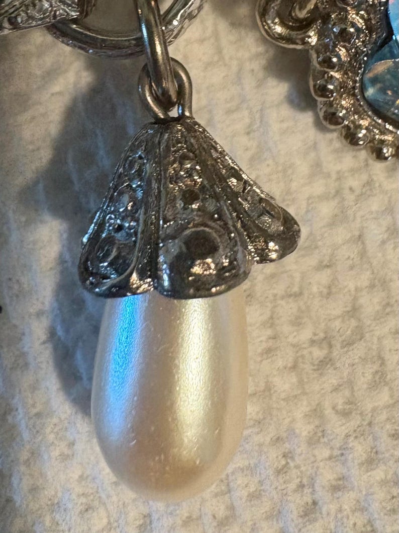 May include: Close-up of a silver-toned earring pendant featuring a pearlescent, teardrop-shaped faux pearl. The pendant is suspended from a decorative, filigree-style cap. The earring is likely vintage or antique.
