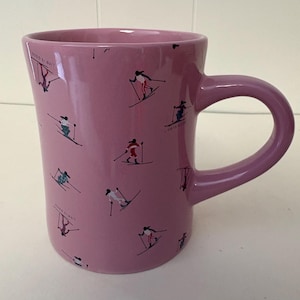 Op de afbeelding: Een mauve keramische mok met een herhalend patroon van kleine figuren die in verschillende richtingen skiën. Het ontwerp bevat kleine illustraties van skiërs in rood, wit en blauwe outfits, samen met de tekst "LIFE IS GOOD" in kleine letters.