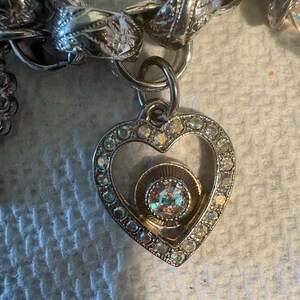 May include: Silver-toned charm bracelet with a heart-shaped pendant. The pendant is encrusted with clear rhinestones around the edges and features a central iridescent stone. The bracelet has a chunky chain design.