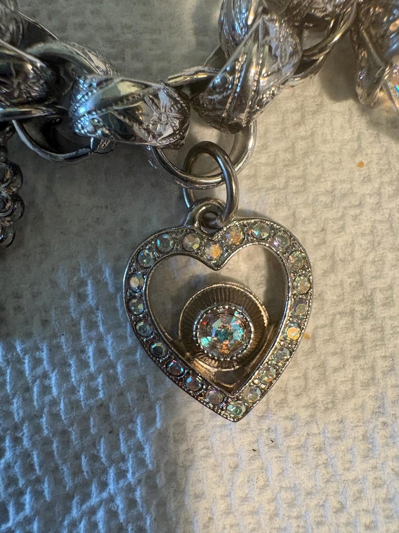 May include: Silver-toned charm bracelet with a heart-shaped pendant. The pendant is encrusted with clear rhinestones around the edges and features a central iridescent stone. The bracelet has a chunky chain design.