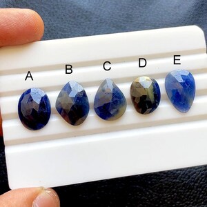 May include: Five faceted blue gemstones, labeled A through E, are displayed on a white surface. The gemstones vary in shape, including oval and teardrop designs. The gems have a deep blue color with reflective facets.