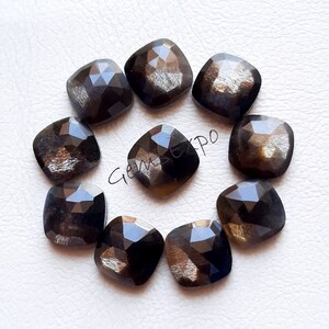 Pode incluir: Uma cole&ccedil;&atilde;o de nove pedras preciosas quadradas e facetadas. As pedras t&ecirc;m uma cor escura e iridescente com tons de dourado e castanho. As pedras est&atilde;o dispostas em um padr&atilde;o circular sobre uma superf&iacute;cie branca. Cada pedra tem cerca de 1,5 cm.