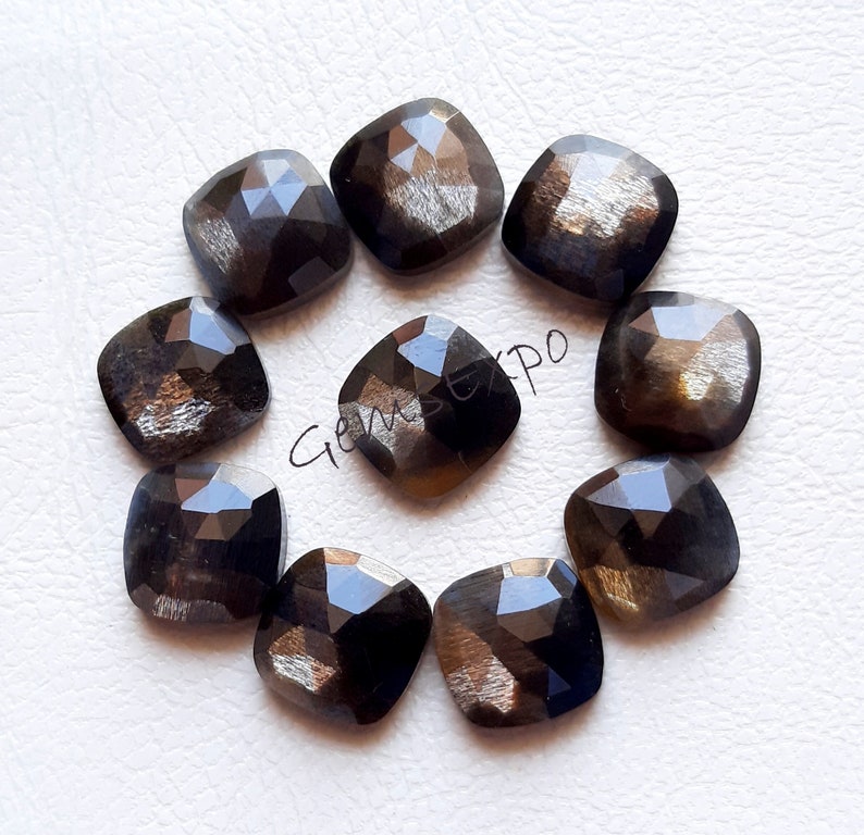 Pode incluir: Uma cole&ccedil;&atilde;o de nove pedras preciosas quadradas e facetadas. As pedras t&ecirc;m uma cor escura e iridescente com tons de dourado e castanho. As pedras est&atilde;o dispostas em um padr&atilde;o circular sobre uma superf&iacute;cie branca. Cada pedra tem cerca de 1,5 cm.