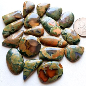May include: A collection of 15 green, brown, and orange polished gemstone cabochons. The cabochons are various shapes and sizes, with some being oval, teardrop, and rectangular. The cabochons are arranged in a pile on a white surface.