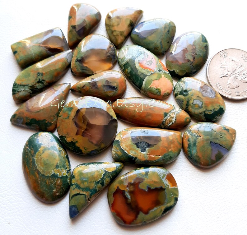 May include: A collection of 15 green, brown, and orange polished gemstone cabochons. The cabochons are various shapes and sizes, with some being oval, teardrop, and rectangular. The cabochons are arranged in a pile on a white surface.