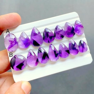May include: A collection of polished purple amethyst gemstones arranged in two rows on a white display card. The stones feature teardrop and oval shapes with visible dark inclusions. The rows are labeled with the letters A and B in black ink.