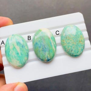 May include: Three oval-shaped gemstones in shades of green and blue are displayed on a white tray. The gemstones are labeled A, B, and C. Each stone has a unique pattern and color variation, with streaks and swirls of different hues.