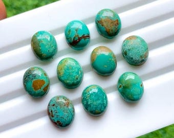 May include: A collection of ten oval-shaped turquoise gemstones. The stones display a vibrant turquoise color with brown and gold matrix patterns. The gemstones are arranged on a white surface, showcasing their polished, smooth surfaces.