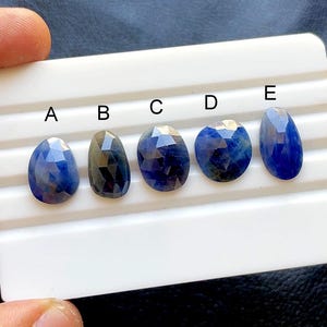 Puede incluir: Cinco zafiros azules facetados de diferentes formas y tamaños se exhiben sobre una superficie blanca. Las piedras preciosas varían en color, desde el índigo intenso hasta tonos de azul más claros, con estructuras internas visibles. Cada piedra está etiquetada con una letra de la A a la E.