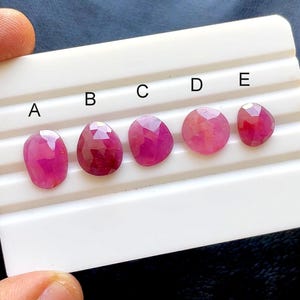 Puede incluir: Cinco gemas facetadas de color rosa-rojo, de forma ovalada, se exhiben en una bandeja blanca. Las gemas están etiquetadas de la A a la E. Las gemas tienen una superficie pulida y reflectante.