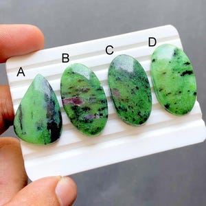 May include: Four polished zoisite gemstones with ruby inclusions, labelled A, B, C, and D. Stone A is pear-shaped, while stones B, C, and D are oval. The stones feature a vibrant green base colour with black speckles and patches of deep red.