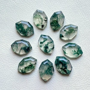 May include: Nine faceted moss agate gemstones with a clear, translucent base and intricate green, tree-like inclusions. The gemstones are arranged on a white surface, showcasing their unique patterns and shapes. Each stone is approximately 1 inch in length.