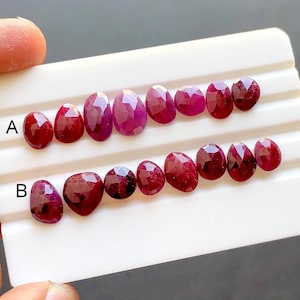 Pode incluir: Uma coleção de rubis vermelho-escuros facetados dispostos em duas fileiras numa bandeja de exibição branca. A fileira superior está marcada com A e a inferior com B. Cada pedra preciosa apresenta uma superfície polida e multifacetada que reflete a luz, exibindo várias formas ovais e de pera em tons carmesim vibrantes.