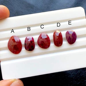 Puede incluir: Cinco piedras preciosas rojas facetadas etiquetadas como A, B, C, D y E, dispuestas en fila sobre una bandeja de presentación blanca con ranuras. Cada piedra presenta una forma irregular única y tonos de rojo intenso con inclusiones visibles.