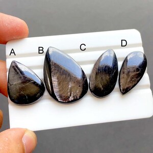 Puede incluir: Cuatro piedras preciosas negras pulidas de varios tamaños se muestran en una bandeja blanca. Las piedras tienen una superficie brillante y reflectante con sutiles patrones internos. Cada piedra tiene forma de lágrima, con una parte superior lisa y redondeada y una base cónica.