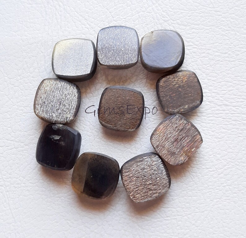Pode incluir: Uma cole&ccedil;&atilde;o de dez pedras preciosas quadradas em v&aacute;rios tons de cinza, marrom e preto. As pedras t&ecirc;m uma forma polida, ligeiramente arredondada e uma superf&iacute;cie texturizada. O texto "GemsExpo" &eacute; vis&iacute;vel.