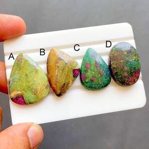 Könnte beinhalten: Vier polierte Cabochon-Edelsteine, beschriftet mit A, B, C und D. Stein A ist hellgrün mit roten Einschlüssen. Stein B ist goldbraun mit roten Flecken. Stein C ist dunkelgrün mit roten Sprenkeln. Stein D ist dunkel, gesprenkelt grün mit roten Flecken.