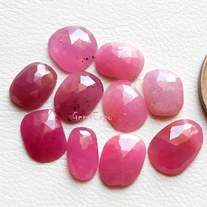 May include: Ten faceted pink ruby gemstones, each with a unique shape and size. The gemstones are arranged in a cluster on a white surface.