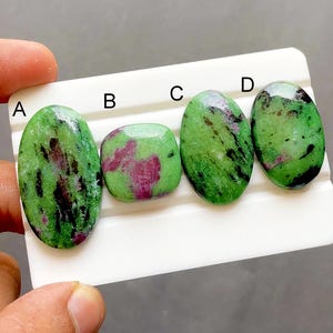 May include: Four polished zoisite gemstones with ruby inclusions, labeled A, B, C, and D on a white display card. The stones feature a vibrant green base with dark black mineral streaks and patches of deep magenta-pink ruby throughout the surfaces.