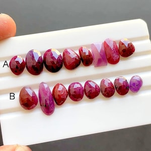 May include: A collection of faceted ruby gemstones in various shapes, including oval and rectangular cuts. The rubies display a deep red hue and are arranged on a white surface. The gemstones are labeled with the letters "A" and "B".