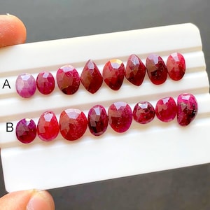 Op de afbeelding: Een assortiment gefacetteerde robijn edelstenen in verschillende vormen, waaronder ovaal en peervormig. De robijnen vertonen een dieprode tint en zijn gerangschikt op een witte tray. De edelstenen zijn gemarkeerd met de letters A en B.