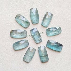 May include: A collection of nine light blue, rectangular gemstones with faceted surfaces. The gemstones are arranged in a circular pattern. The text "GemsExpo" is visible on one of the gemstones.
