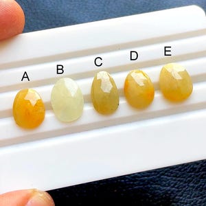 Puede incluir: Cinco piedras preciosas ovaladas y facetadas dispuestas en fila sobre una tarjeta de presentación blanca, etiquetadas de la A a la E. Las piedras varían de color amarillo pálido a naranja dorado intenso, con superficies pulidas y facetas.