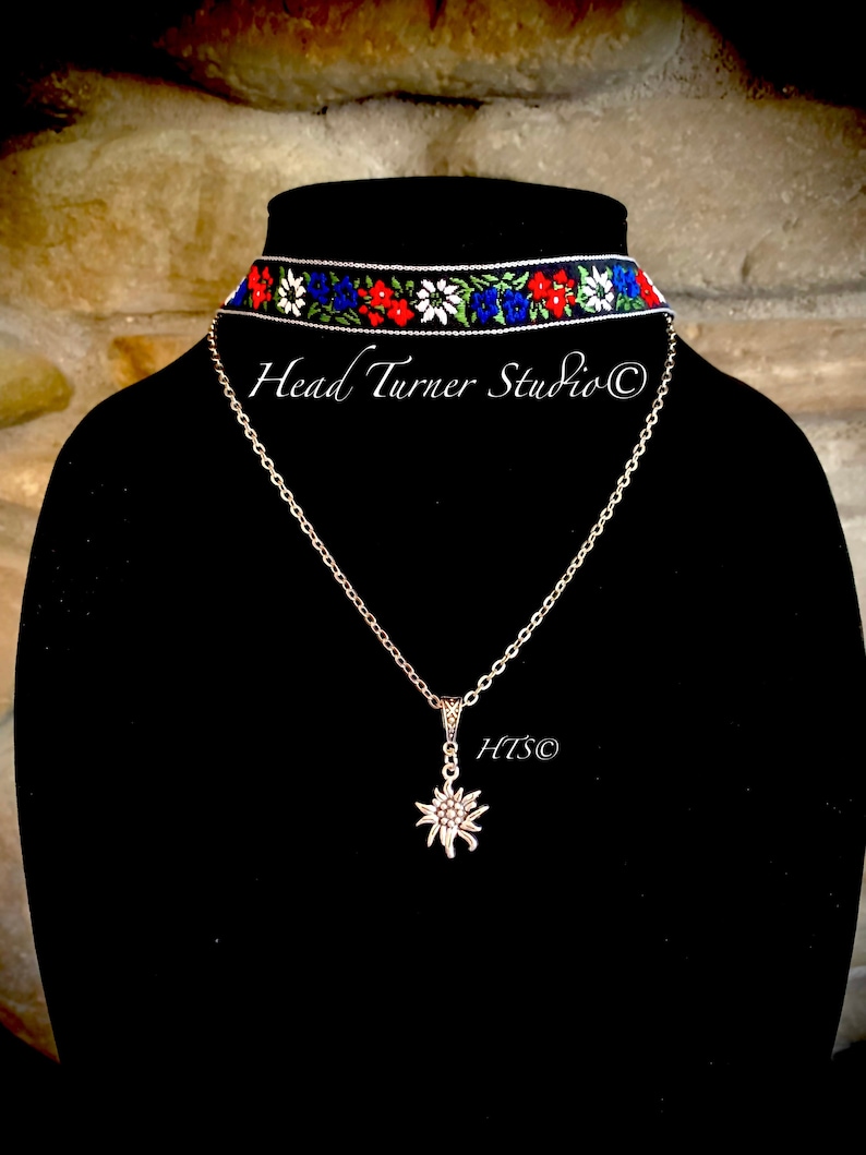 May include: A black choker necklace with a white, red, and blue floral pattern. A silver chain necklace with a silver edelweiss pendant hangs below the choker. The text "Head Turner Studio &copy;" is visible on the black background.