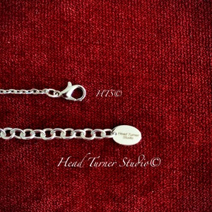 May include: Silver chain necklace with a silver charm that reads "Head Turner Studio" on a red velvet background.