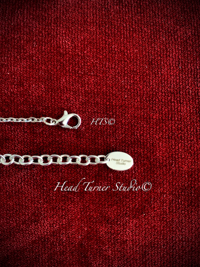 May include: Silver chain necklace with a silver charm that reads "Head Turner Studio" on a red velvet background.